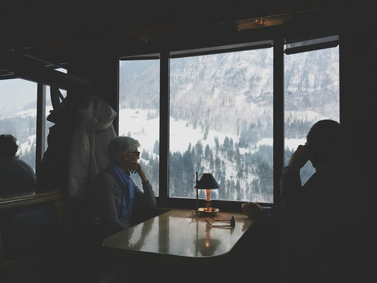 An elderly woman sitting indoors looking out at a snow-covered winter landscape — representing the months when people in the UK stay indoors and sunlight is too weak to produce Vitamin D